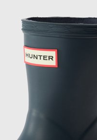 Botte de pluie en caoutchouc bleu foncé avec un bord supérieur texturé et une étiquette rouge et crème bien visible portant la mention "HUNTER". Surface lisse avec une finition mate.