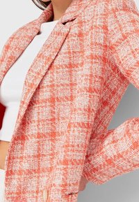 Donna che indossa una giacca a scacchi arancione e bianca sopra un crop top bianco, in piedi con una mano sulla vita.
