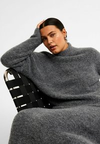 Grå, oversized sweater med høj hals, blødt, fuzzy tekstur og lange ærmer, der hænger over en sort vævet stol.