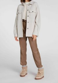 Osoba w kremowej kurtce zapinanej na guziki na białej bluzce, w brązowych spodni high-waist i beżowych zimowych butach na sznurówki z skarpetkami.