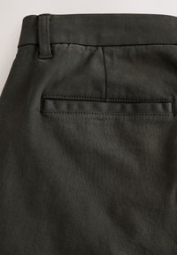 Gros plan d’un pantalon en tissu foncé montrant une poche passepoilée arrière et un passant de ceinture près de la taille.