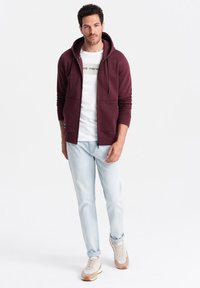 Bordeauxkleurige zip-up hoodie met een voorkrui pocket, gedragen met een wit T-shirt met een bedrukte afbeelding, lichtblauwe jeans en beige sneakers.