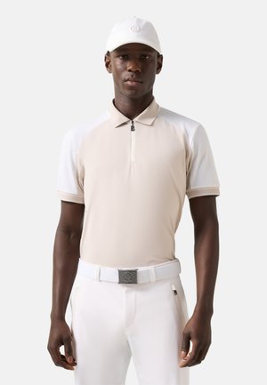 Mand iført beige og hvid kortærmet polo, hvide bukser, hvidt bælte og hvid kasket, stående mod en ensfarvet baggrund.