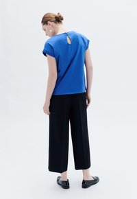 Top blu corto a maniche corte con una scollatura a chiave sul retro, abbinato a pantaloni neri larghi e scarpe nere, presentato da un lato posteriore su uno sfondo chiaro.