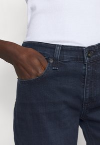 Donkerblauwe spijkerbroek met een front pocket en knoopsluiting. De stof heeft een gladde textuur en subtiele vervaging. Close-up van het taillegebied.