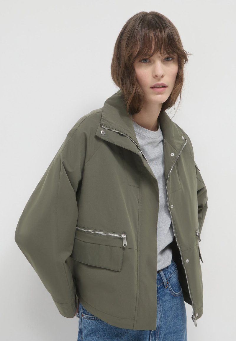 Femme portant une veste zippée vert olive avec des poches à rabat et zippées, un t-shirt gris clair et un jean bleu, sur fond clair.