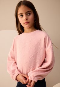 Maglione rosa chiaro con motivo strutturato a fiocchi, maniche lunghe e polsini arricciati. Il tessuto sembra morbido e leggermente spesso.