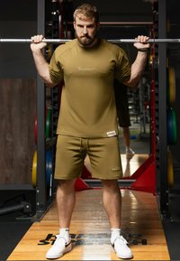 Ensemble sportif vert olive composé d'un t-shirt à manches courtes et d'un short. Les deux présentent une coupe décontractée et un marquage subtil. Porté avec des baskets blanches.