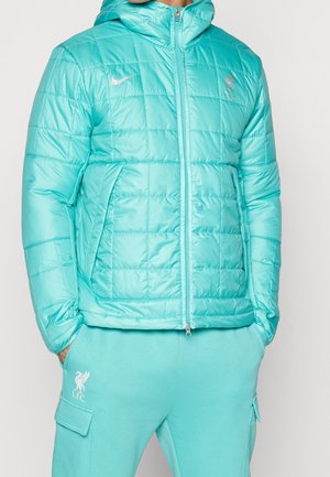 Mężczyzna w jasnoniebieskiej pikowanej kurtce i pasujących spodniach dresowych Liverpool FC, ręce w kieszeniach, stojący na jednolitym tle.