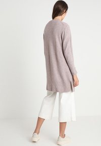 Cardigan long rose clair en matériau doux, présentant une coupe décontractée, des fentes sur les côtés et des poignets côtelés, associé à un pantalon large blanc.