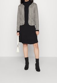 Svart rippad klänning med en kort, mönstrad rutjacka, svarta ankelstövlar och en vit handväska. Jackan har rundade kanter och långa ärmar.