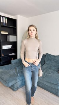 Lys beige, tettsittende langermet topp kombinert med høyt midjer, falmede blå jeans. Hun bærer en liten crossover-veske og står i et koselig rom.