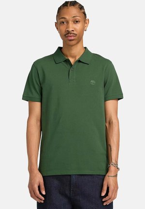 Uomo con i capelli intrecciati che indossa una polo a maniche corte verde scuro e pantaloni scuri, in piedi davanti a uno sfondo semplice.