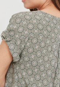 Grøn mønstret bluse med blomster motiver, der har korte, rullede ærmer og en rund halsudskæring. Stoffet har et let, struktureret udseende.