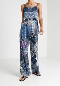 Femme portant un haut à motifs bleu sans manches et un pantalon large assorti, avec des sandales noires, se tenant sur un fond uni.