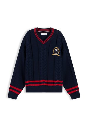 Junge mit lockigem Haar lächelt, trägt einen marineblauen Kabelstrickpullover mit roten Streifen und Emblem, weißes Hemd und hellblaue Jeans.