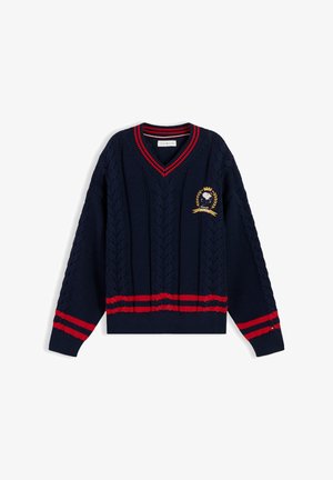 Junge mit lockigem Haar lächelt, trägt einen marineblauen Kabelstrickpullover mit roten Streifen und Emblem, weißes Hemd und hellblaue Jeans.