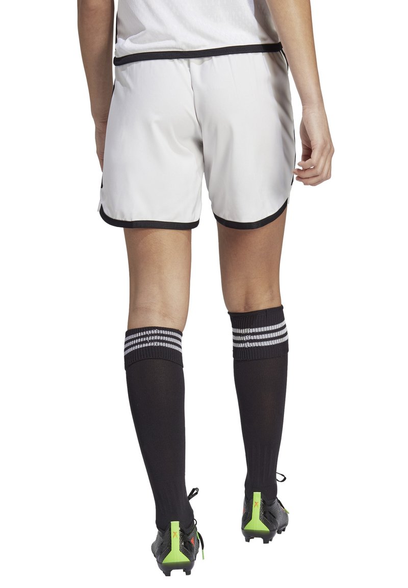 Shorts de sport blancs avec des effets noirs, associés à des chaussettes noires avec des rayures blanches. Vue de dos avec des chaussures de football noires.