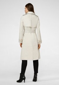 Beige trench coat with a belted waist, shoulder epaulettes, and long sleeves, paired with black ankle boots and fitted black pants.