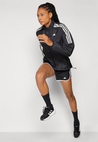 Must värviline spordijakk, millel on valged triibud ja Adidas logo. Kombineeritud mustade lühikeste pükste ja sokkidega, rõhutades sujuvat ja funktsionaalset disaini.