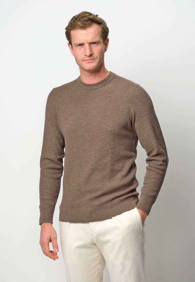 Maglione lungo marrone con texture e maniche lunghe, colletto rotondo, indossato con pantaloni bianchi. Colore neutro, design aderente e polsini a coste.