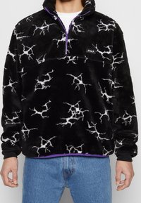Pull noir duveteux avec motifs d'éclairs blancs, fermeture quart de zip violette, logo "HUF" sur la poche poitrine, porté avec un jean bleu et un t-shirt blanc en dessous.