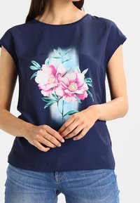 Marinblå kortärmad topp med ett blommönster i rosa pioner och gröna blad, tillverkad av ett glatt tyg. Figursydd siluett med en subtil lyster.