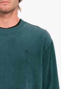 Sudadera en color azul verdoso con cuello redondo, fabricada con un tejido texturizado y un logo en relieve en el lado izquierdo del pecho. Presenta un corte holgado y puños de canalé.