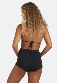 Costumini da bagno neri a vita alta in tessuto testurizzato, abbinati a un semplice bikini nero con allacciatura posteriori. Vista posteriore che mostra l'acconciatura.