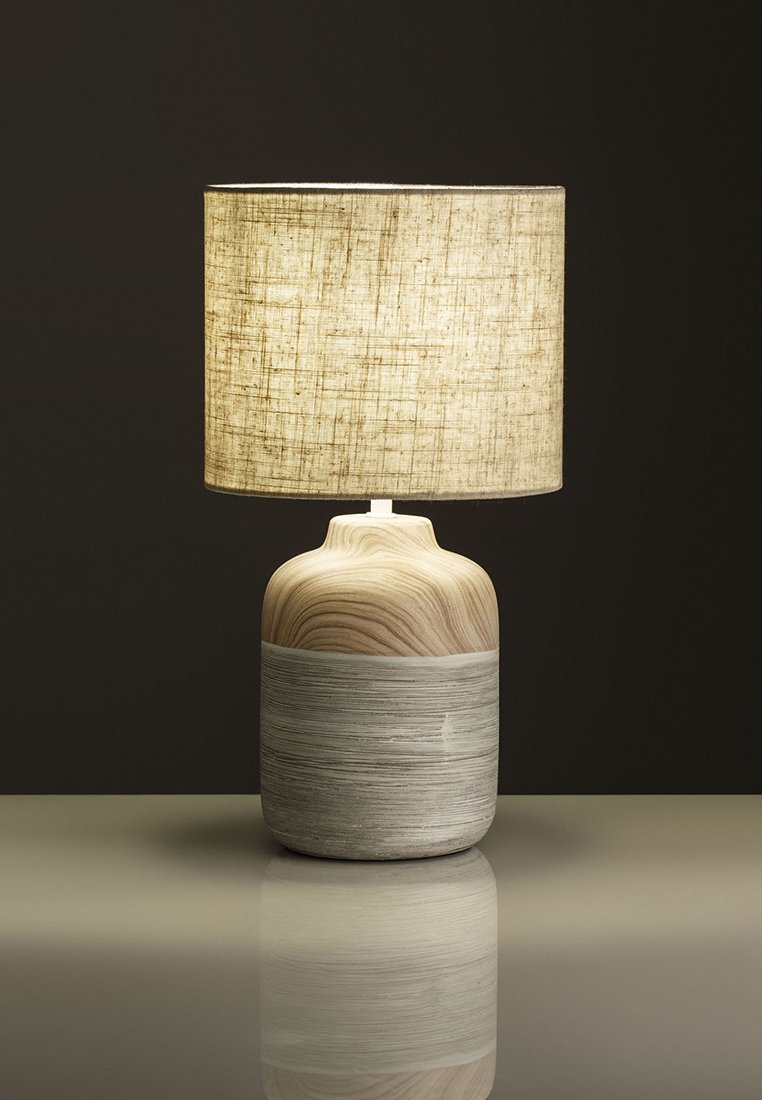 Tischlampe mit einem abgerundeten Holzfuß in hellen und grauen Tönen, gekrönt von einem strukturierten Beigefarbigen Stoffschirm. Reflektierende Oberfläche darunter.