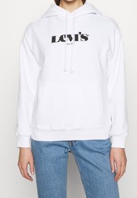 Felpa bianca con tasca frontale e cappuccio regolabile, con il logo nero "Levi's" sul petto. Abbinata a jeans in denim blu.