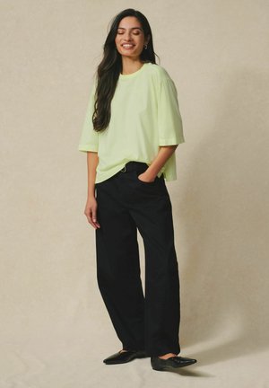 Pitkähiuksinen nainen, joka käyttää vaaleanvihreää ylikokoista T-paitaa, mustia leveälahkeisia housuja ja mustia teräväkärkisiä balleriinoja, seisoo yhdellä kädellä taskussa.