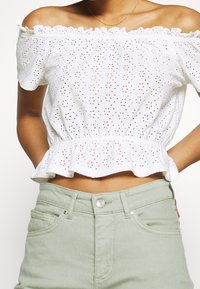 Top blanco sin mangas con patrón de ojal, adornos con volantes y dobladillo de peplum, combinado con pantalones de tiro alto en verde claro.
