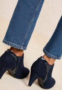 Botines de tacón alto en azul marino, hechos de ante, con un acento de cuero charol y un cremalleras lateral, combinados con unos pantalones de mezclilla de lavado claro.