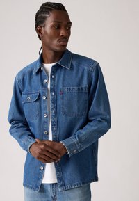 Chemise en denim bleu moyen avec un design à boutons, deux poches poitrine et des coutures renforcées. Dotée de manches longues et d'une coupe décontractée.