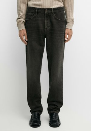 Mand iført falmede sorte jeans, beige ribbet sweater og sorte læder loafers, står mod en ensfarvet hvid baggrund.