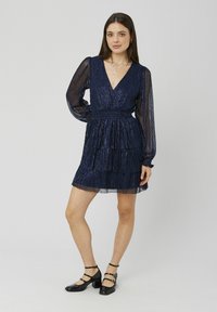 Robe bleu marine avec de longues manches transparentes, décolleté en V profond, jupe à volants et tissu texturé. Associée à des chaussures à talons carrés noires.
