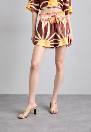 Mannequin portant un short taille haute marron et jaune avec un motif rayons de soleil et des sandales à talons dorées à brides, debout sur un sol gris.