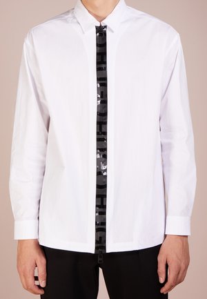 Mies, jolla on päällään valkoinen pitkähiha-paita, jossa on keskellä musta vetoketju ja pystysuoraa kiiltävää tekstiä, yhdistettynä mustiin housuihin.