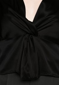 Blusa in satin nero con scollatura a V profonda, dettaglio con nodo intrecciato in vita e tessuto fluente con una texture liscia.