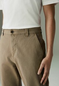 Pantalon en coton marron avec une fermeture à bouton, des passants pour ceinture et des poches latérales. Texture lisse et coupe droite. T-shirt blanc au-dessus.