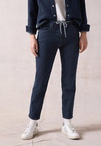 Tmavě modré džíny z denimu s proužkovaným vzorem, s elastickým pasem na šňůrku a bočními kapsami, doplněné bílými teniskami a ponožkami.
