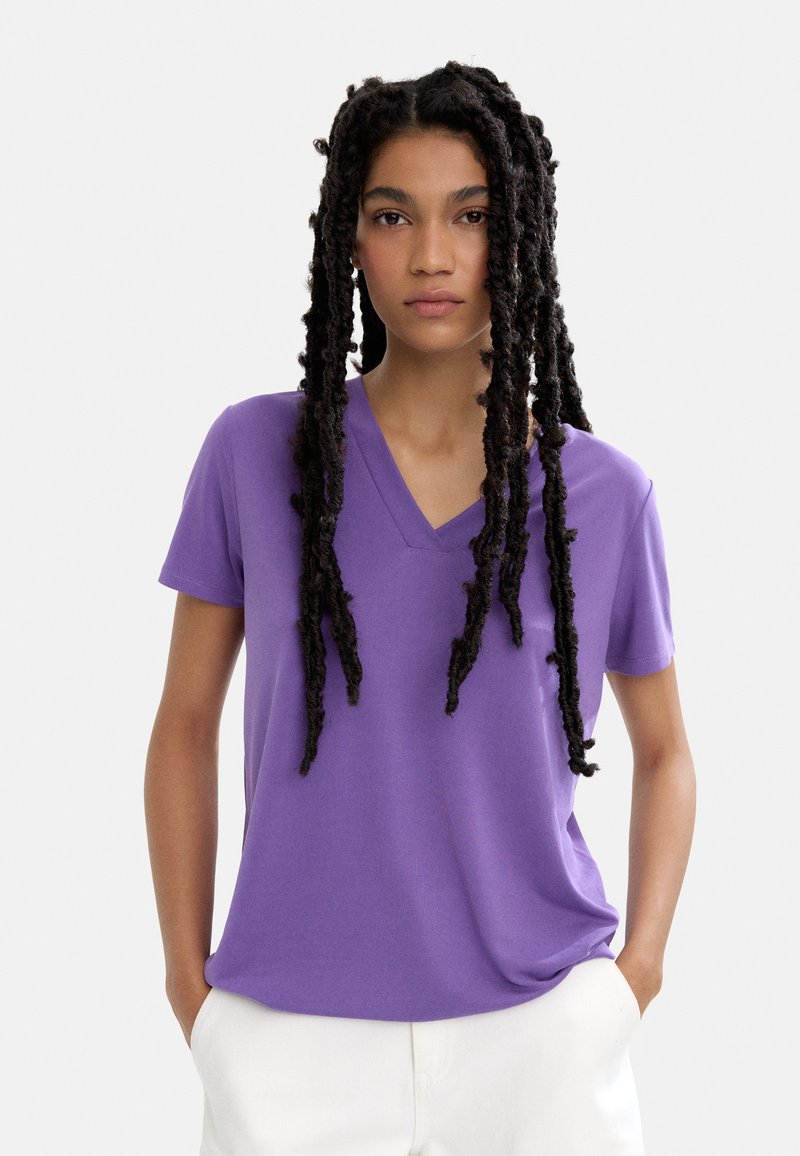 Fioletowa koszulka w serek, wykonana z miękkiego materiału, z krótkim rękawem i luźnym krojem, włożona w białe spodnie o gładkiej fakturze.