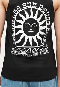 Canotta nera con grafica bianca di un sole stilizzato con volto e testo: "Qui il sole non tramonta mai," "Venti del deserto e cieli infuocati," "Anima dell'ondata di calore - cavalca la luce."