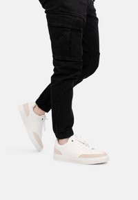 Zapatillas blancas con acentos de ante crema, diseño con cordones y suela de goma gruesa, combinadas con pantalones cargo negros con puños elásticos.