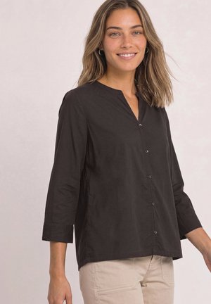 Femme portant une chemise noire à manches longues boutonnée et un pantalon beige, souriant avec les cheveux lâchés sur un fond clair uni.