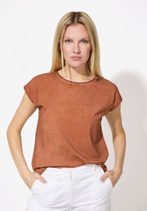 T-shirt couleur rouille, à manches courtes et col rond avec une texture subtile, ornée d'un texte brodé "JE MARCHE SOUS LE SOLEIL" en doré.