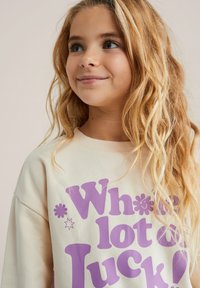Svetlo béžová mikina s fialovým nápisom "Wholesome Luck!". Má mäkkú tkaninu, okrúhly výstrih a voľný strih.