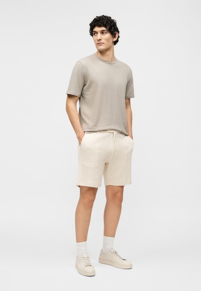 Νεαρός άνδρας στέκεται με τα χέρια στις τσέπες φορώντας μία τσιμέντινη μπλούζα, κρεμ σορτς, λευκές κάλτσες και μπεζ αθλητικά παπούτσια μπροστά σε μια απλή φόντο.