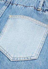 Un bolsillo de denim azul claro con costuras naranjas sobre una tela de denim azul más oscuro, mostrando texturas y detalles de costura.